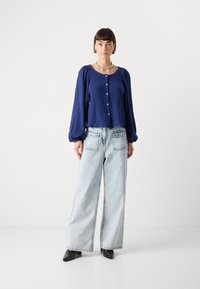 Navy textured button-up top with long puff sleeves, paired with light blue wide-leg jeans and black pointed-toe heels. Neutral background.