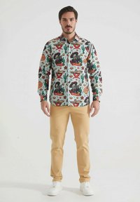 Chemise colorée à manches longues avec un fond clair et divers motifs, y compris des cactus, des maracas et du texte, associée à un pantalon beige.