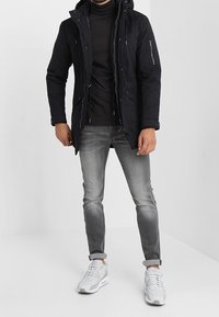 Chaqueta aislante negra con capucha, bolsillos con cremallera y detalles de cremallera, combinada con jeans grises y zapatillas blancas.