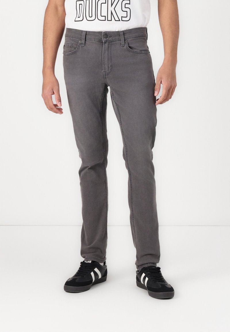 Hombre vistiendo jeans ajustados grises, zapatillas negras con rayas blancas y una camiseta blanca con el texto "DUCKS" parcialmente visible.