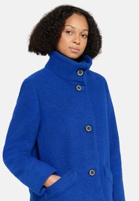 Blauwe wollen jas met een hoge kraag, drie grote knopen en zijzakken. De stof heeft een gestructureerde afwerking. Normale pasvorm.