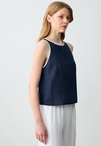 Giovane donna in un top senza maniche di colore blu navy con rifiniture bianche e pantaloni bianchi larghi, in piedi di fronte a uno sfondo chiaro, rivolta a destra.