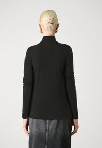 Svart langermet topp med høy krage, glatt tekstur og tettsittende design, kombinert med en grå denimskjørt. Personen ses fra bak.