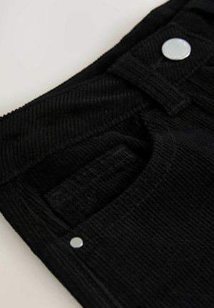 Primo piano di una tasca di pantaloni in velluto a coste neri con dettagli in bottone e rivetti argentati su uno sfondo chiaro.