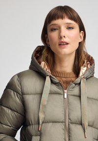 Puffjacka i olivgrönt med dragkedja, huva och kontrasterande bronsofärgad foder. Har dekorativa "POLO CLUB" dragsnören.