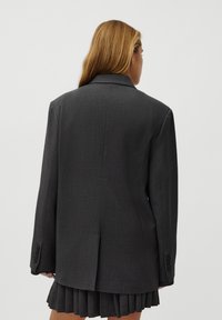 Blazer ajustado em cinza carvão com uma racha única, lapelas em bico e punhos de quatro botões, combinado com uma saia plissada. Textura suave.