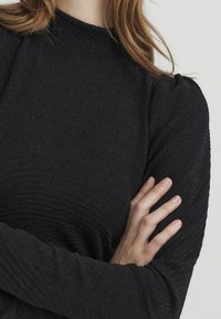 Schwarzes Langarmshirt mit hohem Kragen, ausgestattet mit einem strukturierten Rippmuster und leicht puffigen Ärmeln. Glattes Gewebe mit dezentem Glanz.