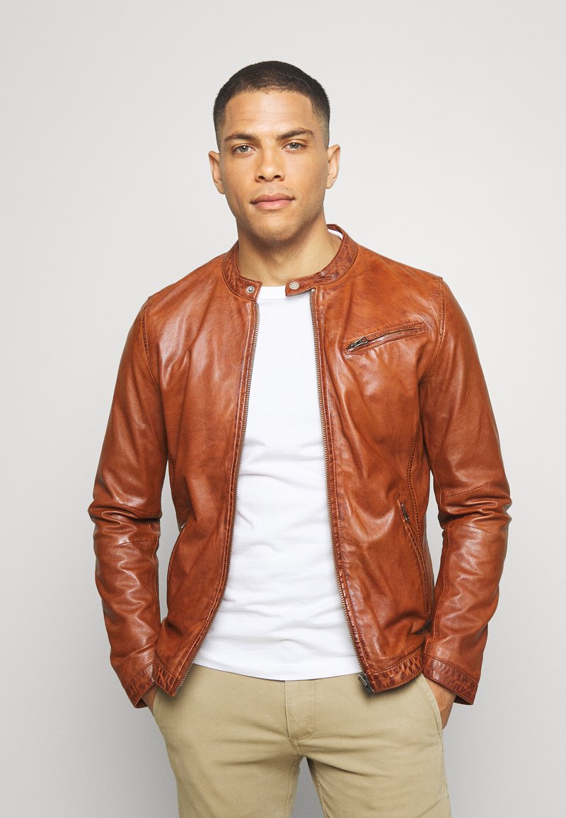 Giacca in pelle marrone con un collo rotondo, zip frontale e tasca laterale con zip. Tessuto liscio e design attillato; indossata sopra una camicia bianca.