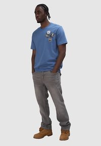 Niebieska bawełniana koszulka z różnymi okrągłymi naszywkami, w połączeniu z szarymi dżinsami i brązowymi roboczymi butami, stojąca z rękami w kieszeniach.