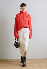 Fleischiger roter Rollkragenpullover mit gerippter Textur, kombiniert mit einem cremefarbenen Midirock mit einem seitlichen Schlitz, schwarzen Stiefeletten und einer kleinen Handtasche.