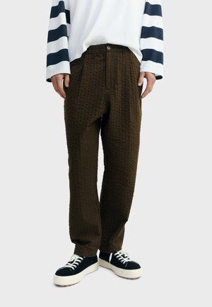 Parte inferior del cuerpo de una persona que lleva pantalones marrón oscuro texturizados, una camisa blanca de manga larga con rayas negras y zapatillas negras con suelas blancas.