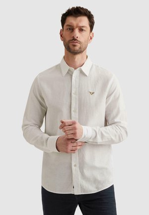 Mężczyzna ubrany w białą, zapinaną na guziki koszulę z długim rękawem, z małym złotym logo orła na lewej piersi, stojący na jednolitym tle.