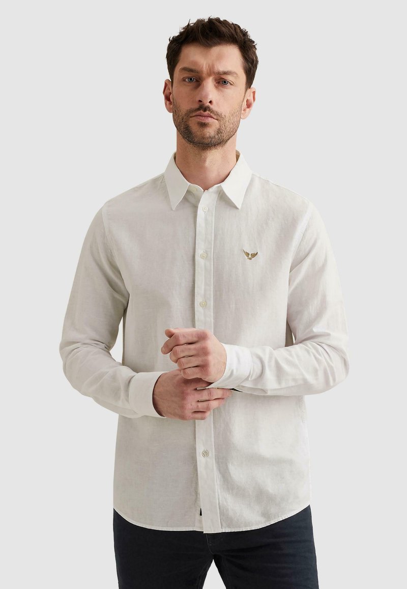 Homme portant une chemise blanche à manches longues boutonnée avec un petit logo aigle doré sur la poitrine gauche, debout devant un fond uni.