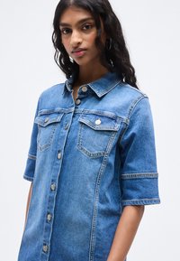 Πουκάμισο από denim σε ανοιχτό μπλε χρώμα με εμπρόσθιο κλείσιμο με κουμπιά, κοντά μανίκια, δύο τσέπες στο στήθος και κλασικό γιακά. Διαθέτει διακριτικές ραφές.