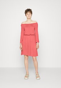 Vestido coral com ombros à mostra, mangas longas, cintura ajustada e saia fluida, feito de tecido macio. Combinado com sandálias de plataforma leves.