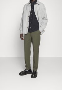 Mann in grauem Überhemd über einem schwarzen Hemd mit Knöpfen, olivgrüner Hose und schwarzen Lederloafers mit weißen Socken vor einem einfarbigen Hintergrund.