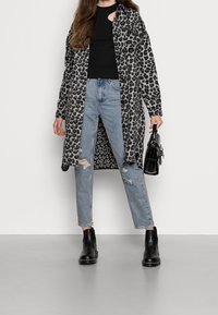 Langarm-Leopardenmuster-Mantel in Grau und Schwarz, kombiniert mit einem schwarzen Oberteil, ausgewaschenen blauen Jeans und schwarzen Ankle Boots. Eine kleine schwarze Handtasche wird gehalten.