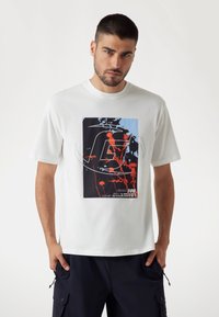 Camiseta blanca de algodón con una gran impresión gráfica que incluye elementos en rojo y azul, como un logo estilizado y patrones abstractos.