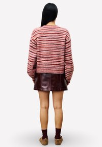 Femme aux longs cheveux noirs portant un pull rayé rouge et rose, une jupe en cuir rouge sombre, des chaussettes bordeaux et des chaussons en peluche marron, de dos.