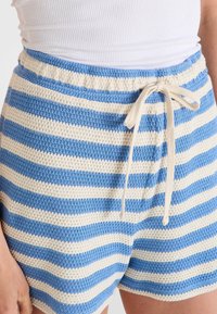 Pantalones cortos a rayas azul y crema con cintura ajustable, parcialmente visibles, combinados con una camiseta blanca de canalé. Atuendo casual de verano.