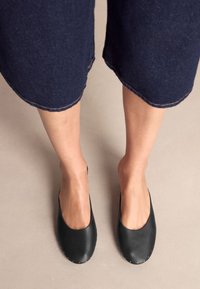 Schwarze Leder-Slipper mit abgerundeten Zehen, gesichert mit goldfarbenen Nieten entlang der Sohle. Kombiniert mit dunkelblauen Jeans-Culottes.