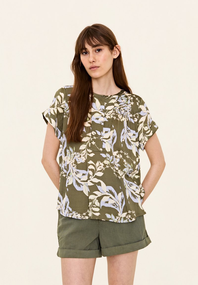Blusa floreale a maniche corte verde con motivi di foglie blu e crema, dotata di scollatura rotonda e chiusura con bottoni. Abbinata a pantaloni corti arrotolati.
