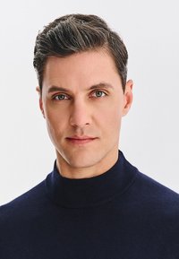 Męski model z krótkimi ciemnymi włosami, ubrany w ciemnogranatowy golf. Tło jest jednolicie białe, co podkreśla rysy twarzy i wyraz modela.