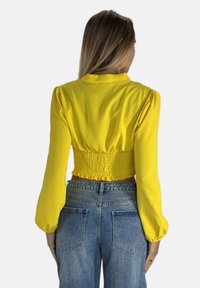 Gul kort blus med puffärmar, smockad rygg och en mjuk textur. Matchad med ljusblå högmidjade jeans.