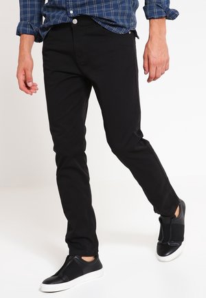 Homme portant un pantalon slim noir, des baskets noires sans lacets et une chemise bleue à carreaux à manches longues, marchant devant un fond blanc.