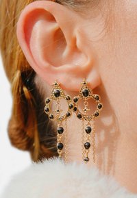Boucles d'oreilles en or avec un design circulaire mettant en avant des pierres noires et des chaînes suspendues. Les détails comprennent de petites étoiles en accent et des sertissages texturés.