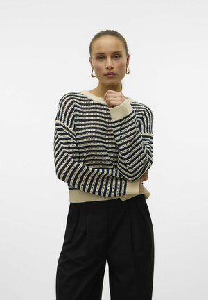 Pull en tricot rayé dans les tons crème, bleu marine et beige, avec une coupe ample, des épaules tombantes et des poignets côtelés, associé à un pantalon noir.