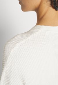 Gros plan sur le cou et l'épaule d'une personne portant un pull en maille blanche texturée, avec des cheveux tressés visibles sur le bord.