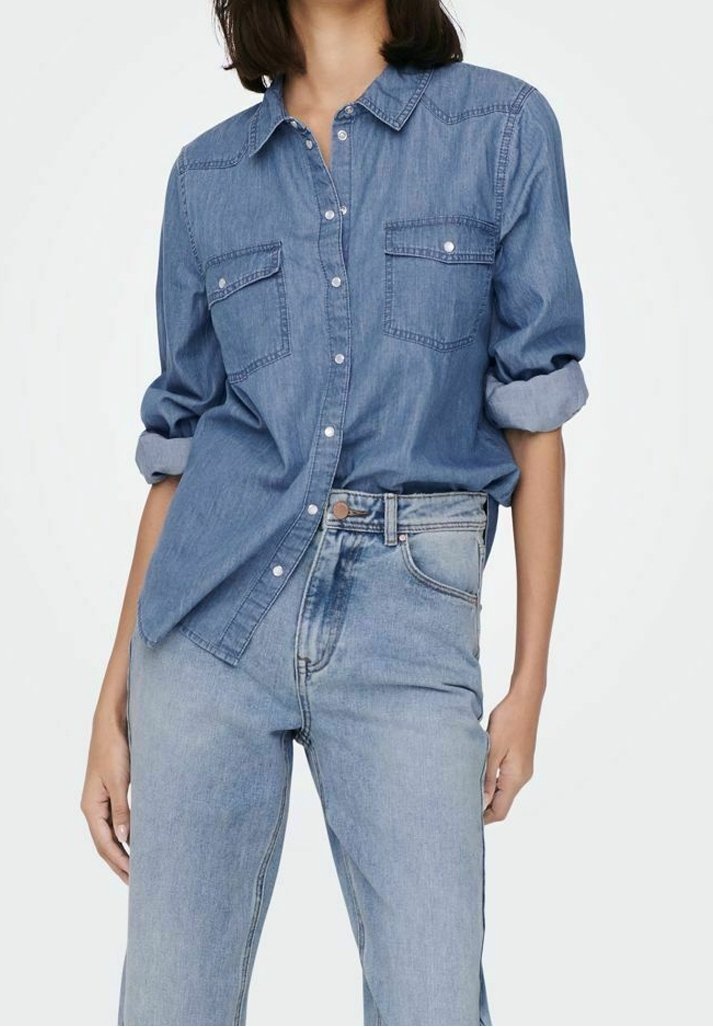 Personne portant une chemise en denim aux manches retroussées, rentrée dans un jean taille haute bleu clair, sur un fond uni.