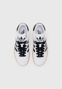 White athletic shoes with black stripes and laces, textured leather upper, and gum rubber outsole. Features iconic logo on tongue.