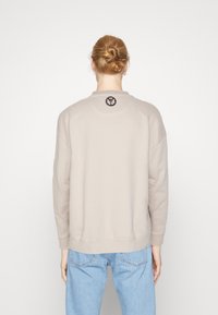 Hellbeige Sweatshirt mit rundem Halsausschnitt und überschnittenen Schultern, mit einem aufgenähten kreisförmigen Emblem auf dem Rücken und gerippten Bündchen.