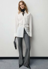 Bijela prevelika košulja s gumbima, sivi trapezni hlače, crne cipele s šiljastim vrhom i mala crna torbica. Glatka tkanina s klasičnim krojem.