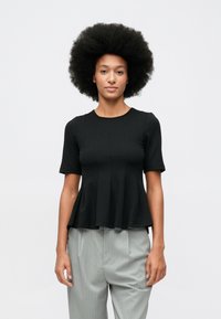 Młoda osoba z bujną afro fryzurą, ubrana w czarną bluzkę peplum z krótkim rękawem i jasnoszare spodnie w prążki, stoi na tle białej ściany.