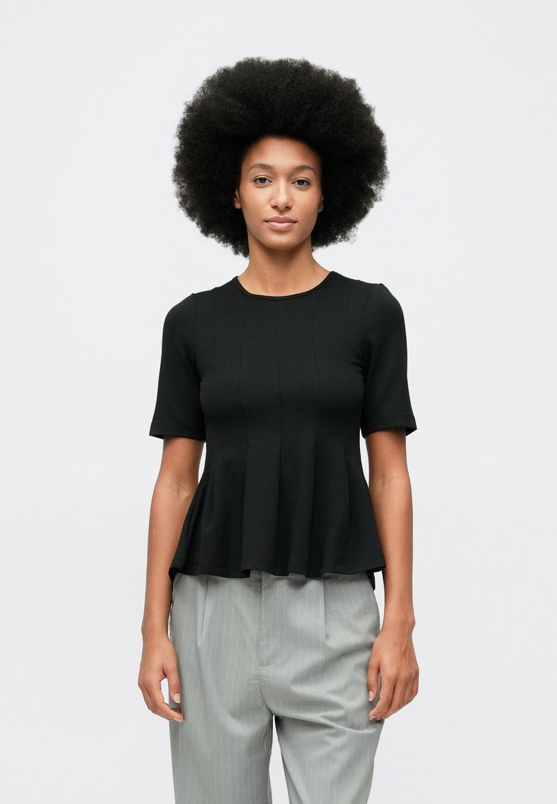 Młoda osoba z bujną afro fryzurą, ubrana w czarną bluzkę peplum z krótkim rękawem i jasnoszare spodnie w prążki, stoi na tle białej ściany.