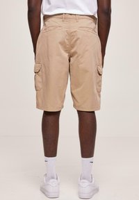 Shorts cargo beiges avec poches latérales, fabriqués en tissu durable, présentant une coupe droite et des coutures visibles au niveau de l'ourlet. Portés avec des baskets blanches.
