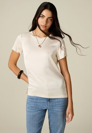 Junge Frau mit langen dunklen Haaren trägt ein cremefarbenes Kurzarmshirt, blaue Jeans, ein schwarzes Armband und eine goldene Anhänger-Halskette vor einfachem Hintergrund.