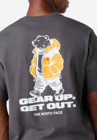 Bjørn i orange jakke og sneakers går, med teksten "Gør dig klar. Kom ud." og "The North Face" på mørkegrå skjorte ryg.