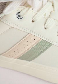 Zapatilla de piel blanca con una franja de acento beige, detalle texturizado verde y secciones perforadas. Incluye cordones redondos y detalle del logo.