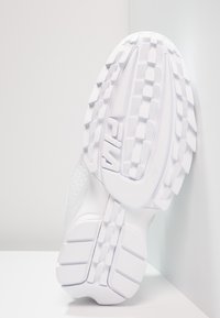 Baskets blanches avec une semelle texturée et surélevée. Elles présentent un dessus lisse avec des perforations et un logo visible sur le côté.