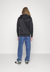 Černá mikina s kapucí s hladkou texturou, žebrovanými manžetami a volným střihem, kombinovaná s volnými modrými džíny a černými teniskami.