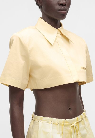 Personne à la peau foncée portant une chemise courte à manches courtes jaune pâle et un pantalon taille haute assorti avec un cordon de serrage.