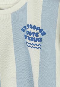 Tissu à rayures verticales bleues et blanches avec texte bleu "St. Tropez Côte d'Azur" et symboles de vagues imprimés sur la zone de la poitrine.