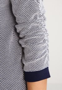 Mörkblå och vit mönstrad stickad tröja med en strukturerad yta och ribbade ärmslut. Har gathered tyg längs armen för extra detalj.