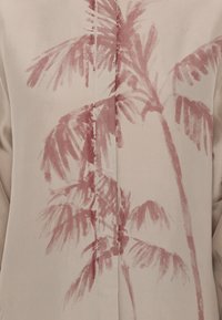 Blusa beige con stampa di foglie di palma in rosso tenue, con una texture liscia e una patta centrale con bottoni che valorizza il design.