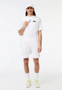 Balta polo krekls ar zaļu krokodila logo, baltas šortas, balta cepure un baltas sporta kurpes ar neona akcentiem. Gludas un plakanas tekstūras.