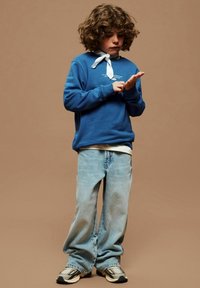 Enfant aux cheveux bouclés portant un sweat-shirt bleu, un jean clair, des baskets et un foulard blanc autour du cou, examinant sa main devant un fond marron uni.